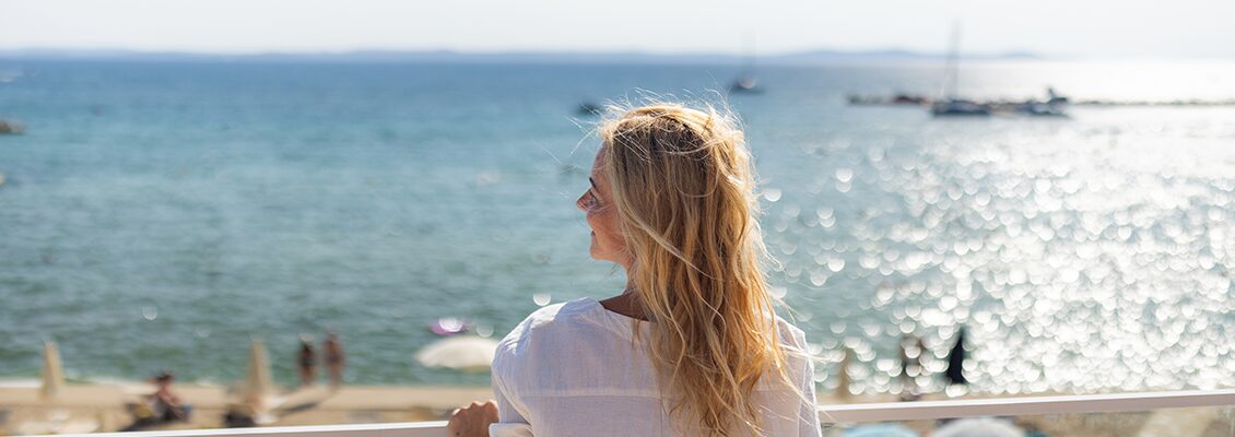 Mulher de cabelos longos e loiros aprecia a vista do mar em um cenário ensolarado e tranquilo, perfeita para momentos de relaxamento na praia.