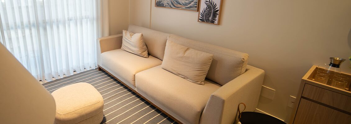 Sofá bege de três lugares em uma sala moderna com decoração minimalista, cortina branca e quadros decorativos na parede, ideal para conforto e elegância.