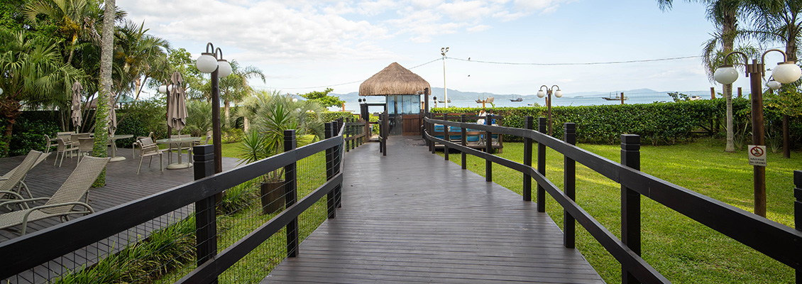Passeio de madeira na orla de praia com gazebo, cercado por árvores e iluminação, com vista para o mar ao fundo, ideal para momentos de lazer ao ar livre.