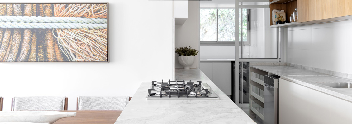 Imagem de uma cozinha moderna com bancada em mármore branco, fogão de quatro bocas e armários brancos, ambiente bem iluminado com janela ao fundo.
