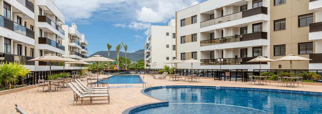 Imagem de uma piscina em um condomínio residencial com áreas de lazer ao redor, cadeiras e guarda-sóis sob um céu azul com nuvens, cercada por prédios modernos.