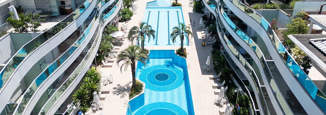 Imagem de uma piscina em um condomínio residencial com áreas de lazer ao redor, cadeiras e guarda-sóis sob um céu azul com nuvens, cercada por prédios modernos.