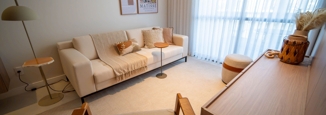 Sala de estar decorada com sofá bege, mesa de centro, poltrona, cortinas transparentes e objetos decorativos em tom neutro, criando um ambiente aconchegante e elegante.