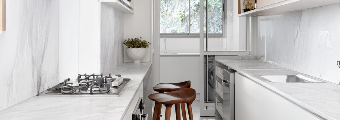 Descrição de uma cozinha moderna e minimalista com balcões brancos, fogão a gás, janelas com vista para o exterior, e duas banquetas de madeira.