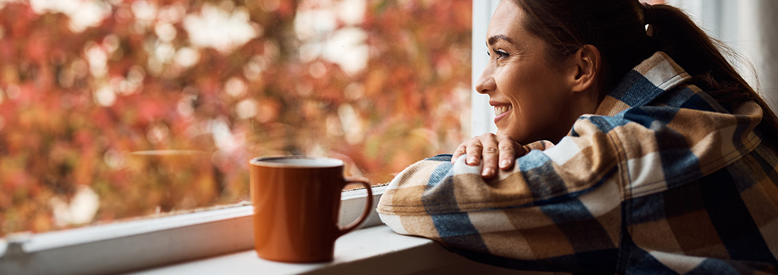 Jovem mulher sorridente apreciando uma xícara de café ou chá enquanto olha pela janela em um ambiente confortável, com fundos de árvores com cores de outono.