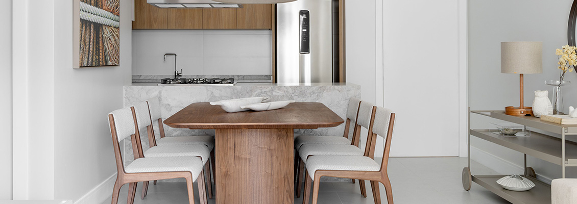 Sala de jantar moderna integrada à cozinha, com mesa retangular de madeira e quatro cadeiras claras, bancada em pedra branca e armários embutidos, fogão ao fundo e decoração neutra. Ambiente sofisticado.