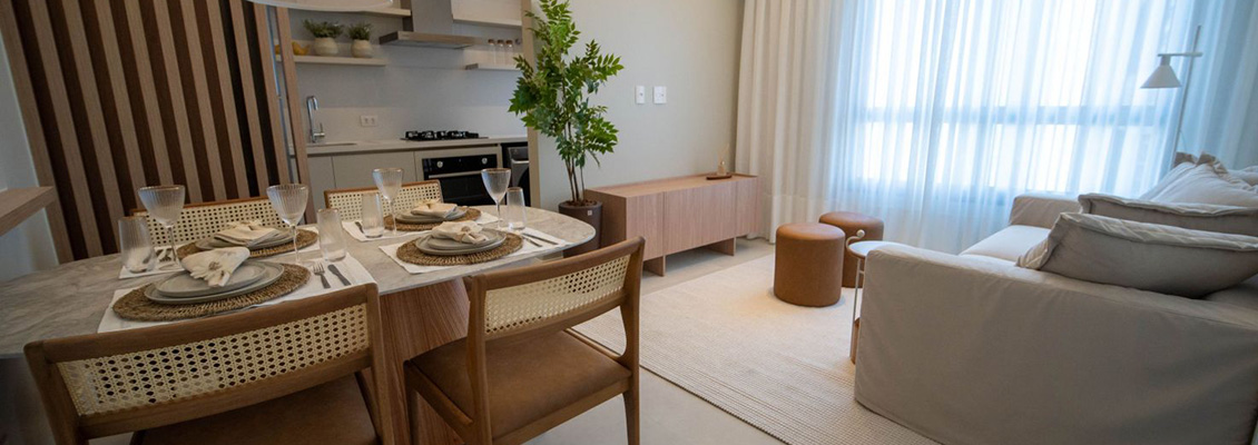 Sala de estar e jantar integrada, com mesa posta para quatro pessoas, cadeiras de madeira e tecido com padrão vazado, cozinha ao fundo e sofá claro ao lado de grandes janelas com cortinas.