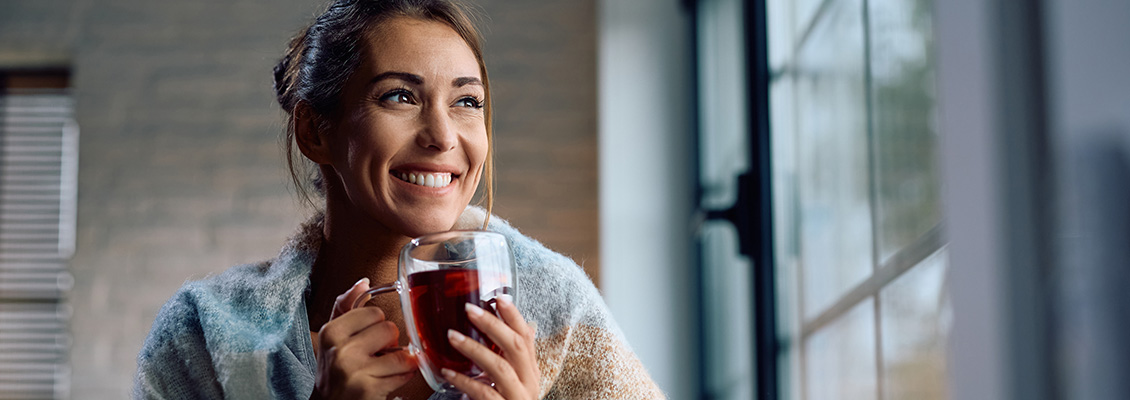 Mulher sorridente em ambiente interno segurando uma caneca com bebida quente, usando roupa casual e com luz natural ao fundo, em clima aconchegante.