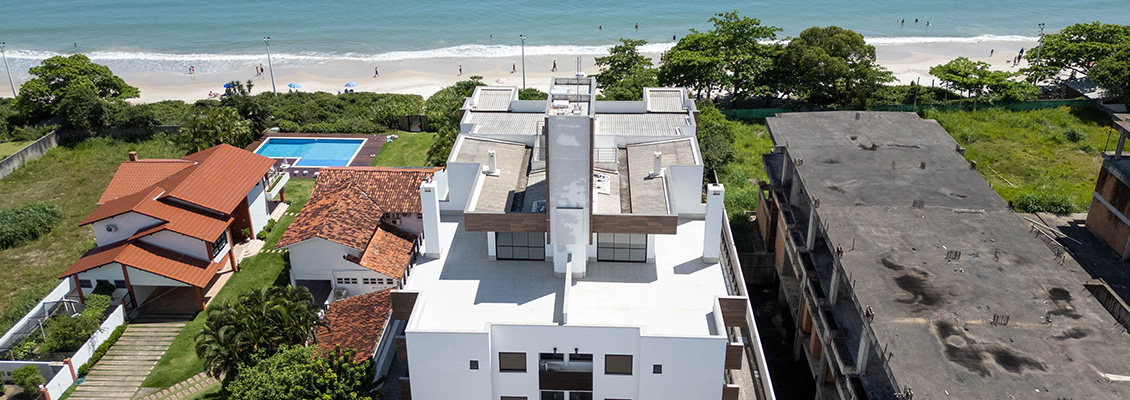 Vista aérea de uma propriedade com casas modernas, telhados em telha e área de lazer com piscina e jardim, ao fundo a praia e o mar.