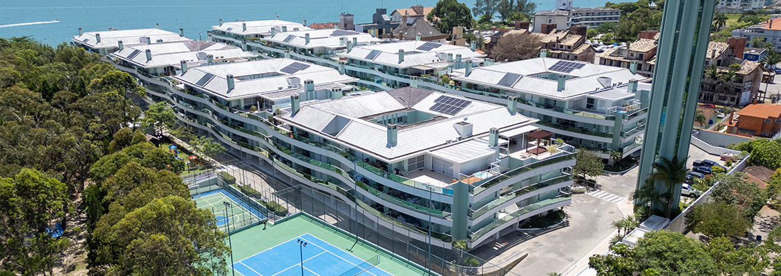 Vista aérea de um condomínio ou complexo residencial com piscinas e quadras esportivas, telhados com painéis solares e prédios de vários andares ao lado do mar