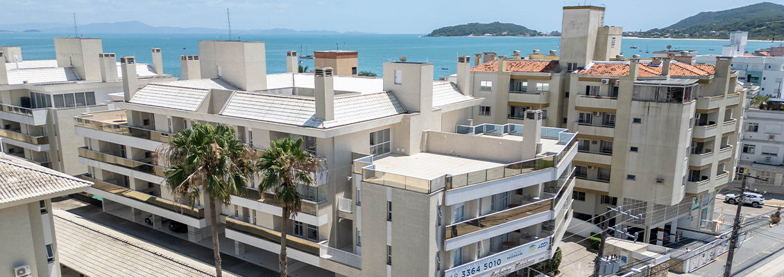 Vista aérea de um conjunto de prédios residenciais com varandas e telhados, cercado por vegetação e mar ao fundo, em Florianópolis