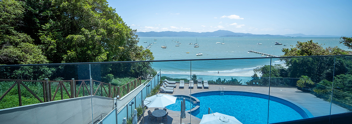 Vista panorâmica de uma piscina ao ar livre com espreguiçadeiras cercada por vegetação verde, olhando para o mar azul e barcos ao fundo