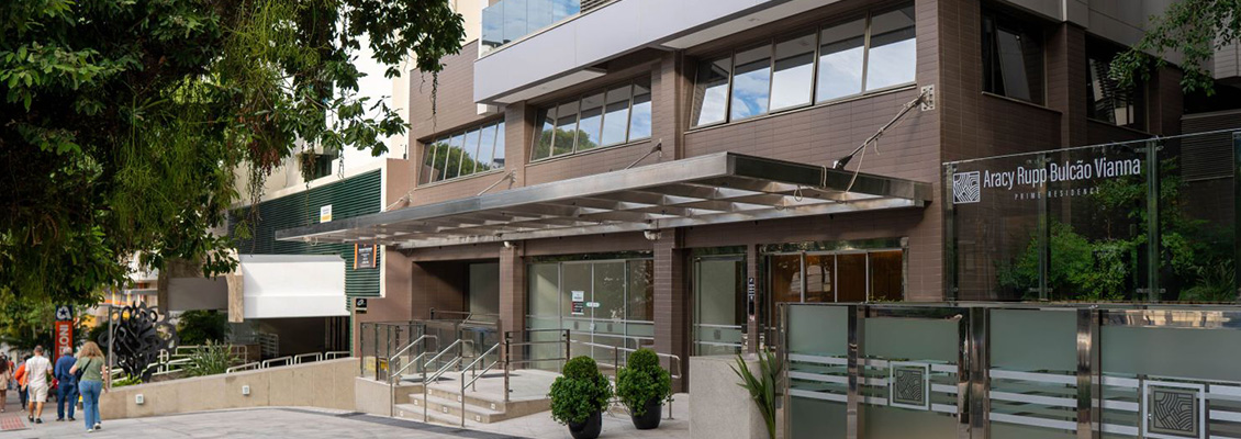 Entrada de prédio corporativo com fachada em vidro e tijolos, marquise com toldo retrátil, rampas de acesso e jardim com árvores ao lado, em ambiente urbano.