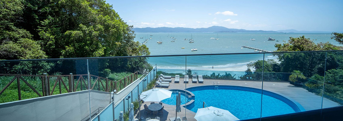 Vista panorâmica de um deck com piscina ao lado do mar, cadeiras ao ar livre e barcos na baía, cercado por vegetação e ao fundo uma linha de montanhas e céu azul.