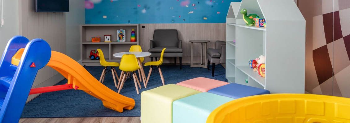 Brinquedoteca infantil com escorregador azul e amarelo, piscina de bolinhas e área com cadeiras e mesa ao fundo, ambiente colorido e seguro.
