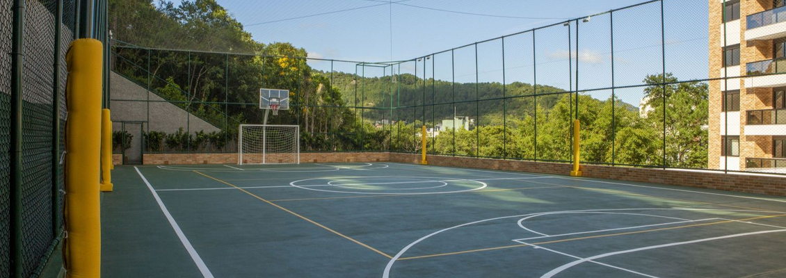 Quadra esportiva ao ar livre com piso azul e demarcações brancas, telas de proteção laterais e goleiras em um complexo residencial.